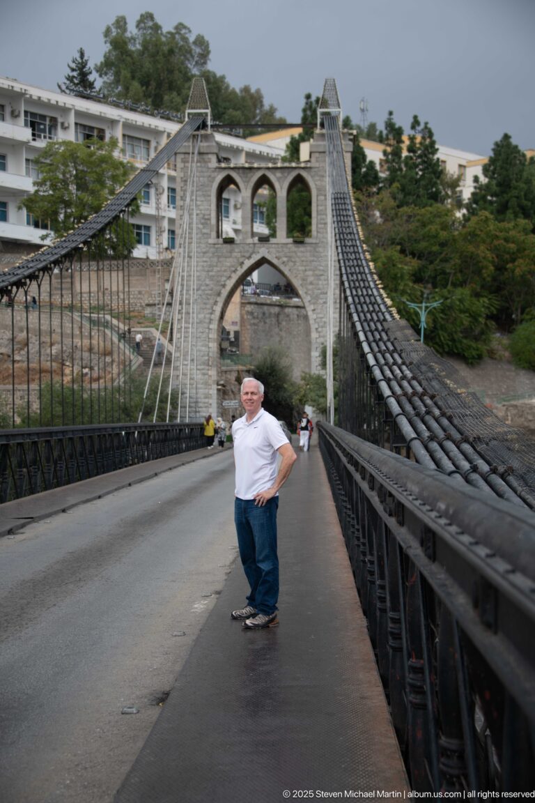 Man standing on Sidi M’Cid Bridge beside heavy suspension cables, with the pointed stone tower beyond.