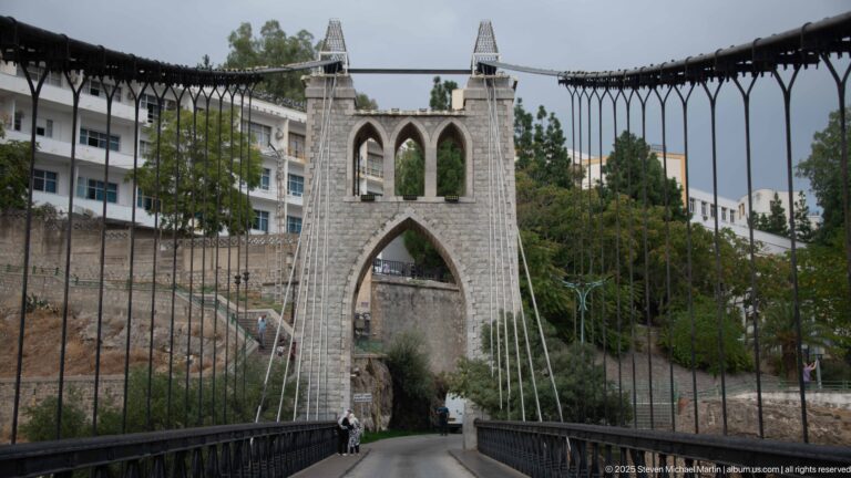 Bridge roadway leading toward Sidi M’Cid’s pointed stone tower, flanked by vertical suspension hangers.