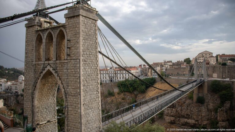 Side view of Sidi M’Cid Bridge tower, suspension cables, deck, and city buildings across Constantine’s gorge.