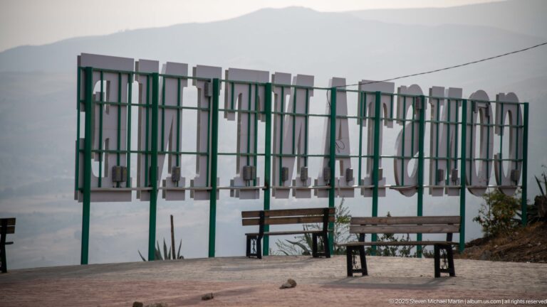 Rear view of large city letters on green metal supports, with benches and hazy hills beyond.