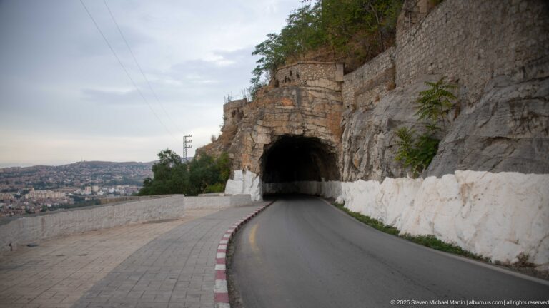 Road entering a rock-cut tunnel along Constantine’s cliff, with whitewashed walls and city buildings in the distance.
