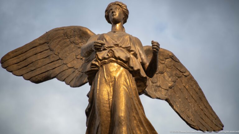 Winged female victory figure with long drapery and raised hands atop Constantine’s Monument aux Morts.