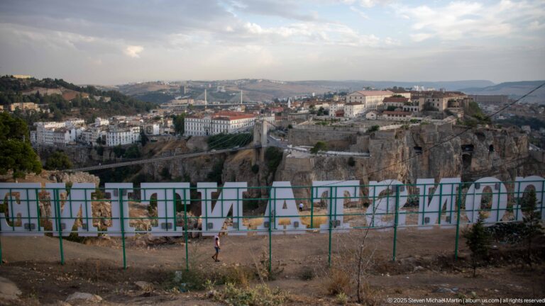 Rear of large CONSTANTINE letters on green supports overlooking the city, gorge, bridges, and distant hills.