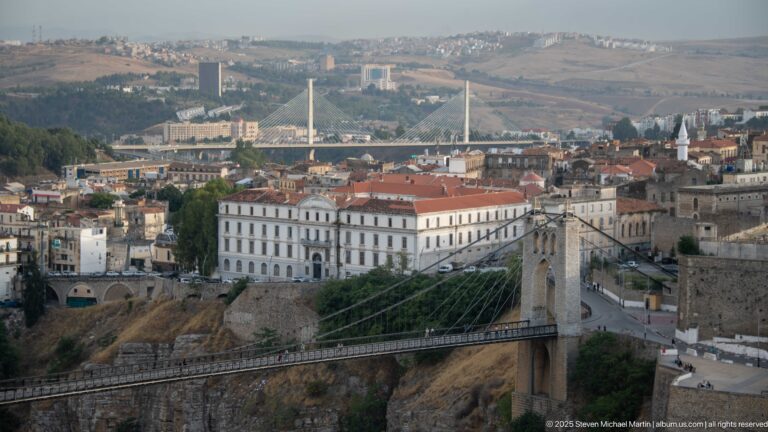 Suspension bridge with stone tower and cables crossing Constantine’s gorge below dense city buildings and distant bridges.