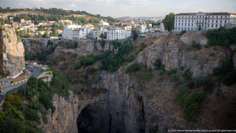 Buildings on the rim of Constantine’s steep gorge above a dark rock opening and curving cliff road.