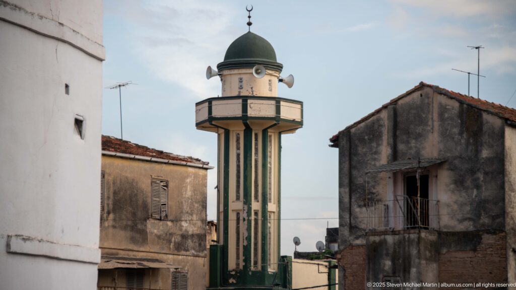 Slender minaret with balcony, loudspeakers, dark dome, and crescent finial rising between Constantine buildings.