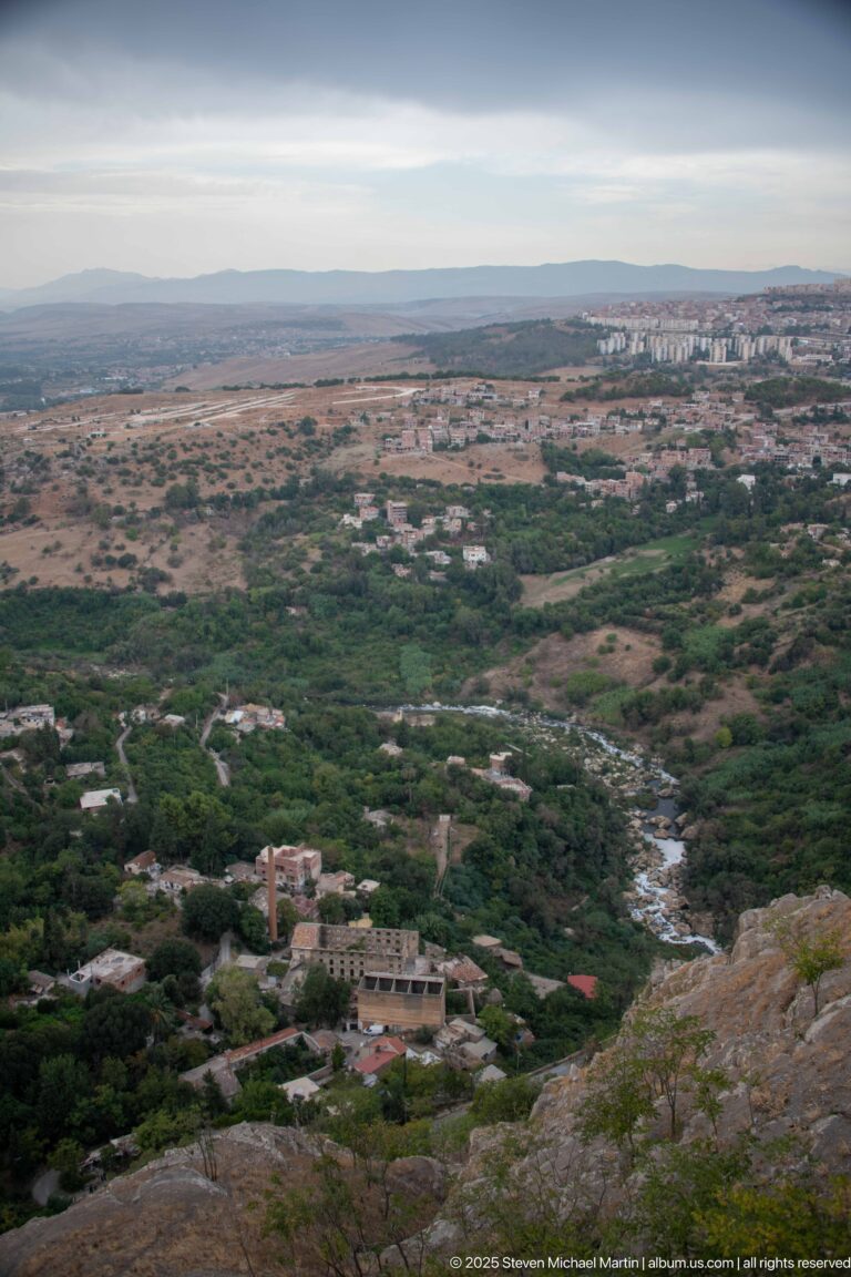 Elevated view over a green valley, scattered buildings, and a narrow watercourse beyond Constantine’s rocky edge.