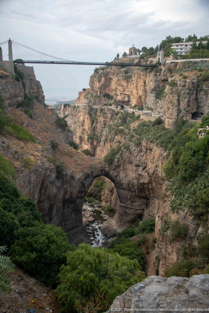 Natural rock arch spanning the Rhumel River at the bottom of Constantine’s gorge, with cliffs and a distant bridge.