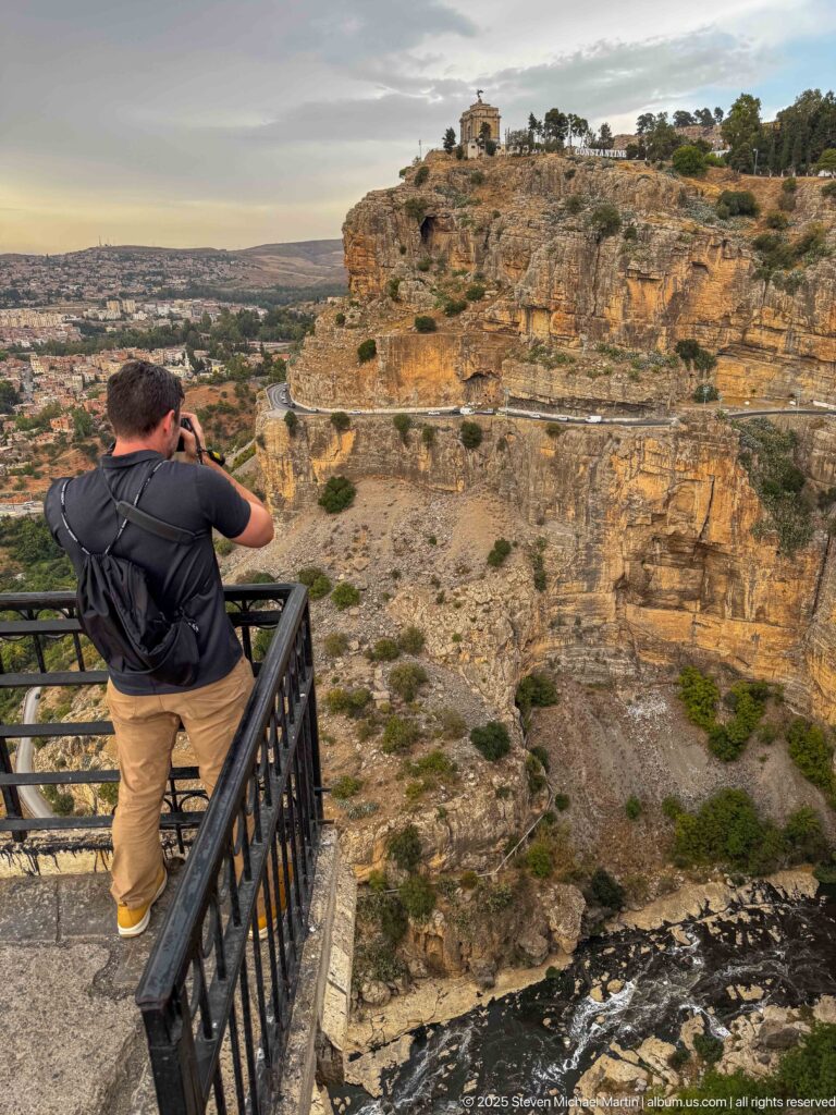 Man photographing a layered cliff across Constantine’s gorge, with a road and Monument aux Morts on the summit.