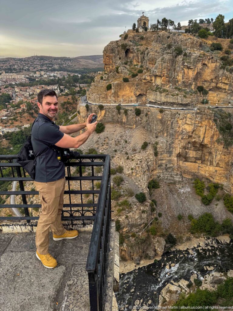 Man holding a phone at a gorge overlook with Constantine’s layered cliffs and hilltop memorial behind him.