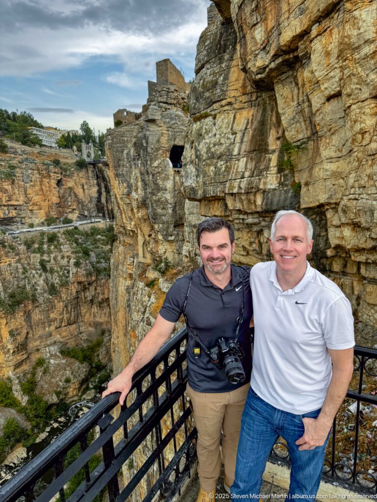 Two men posing at a metal railing beside fractured rock walls in Constantine’s gorge.