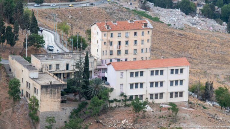 Cluster of pale buildings with red roofs beside a winding road on Constantine’s steep rocky slope.