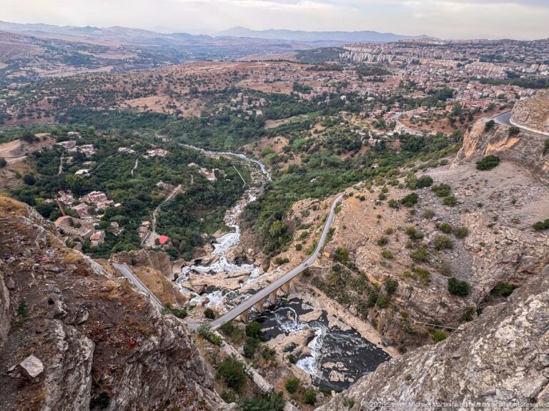 High view down Constantine’s gorge to the Rhumel River, a narrow bridge, road, trees, and distant uplands.
