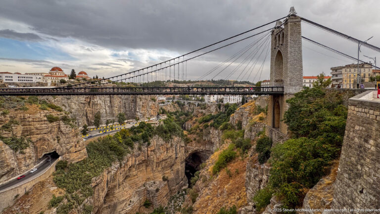 Wide side view of Sidi M’Cid Bridge with stone tower, suspension cables, roadway, cliffs, and city buildings.