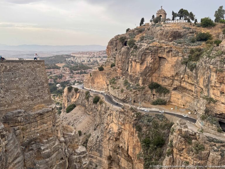 Cliff road below Constantine’s Monument aux Morts and city-name letters, with the gorge and distant neighborhoods beyond.