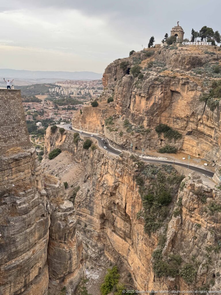 Layered Constantine cliff crossed by a road, with Monument aux Morts and city-name letters on the summit.