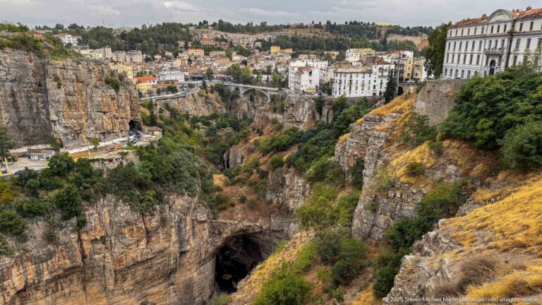 Cliff-top buildings above Constantine’s deep gorge, with a dark rock opening, road, and small bridge below.