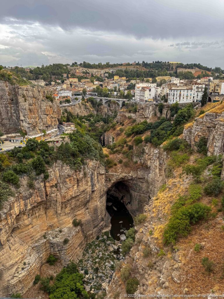 Natural rock arch over the Rhumel River beneath Constantine’s stratified cliffs and cliff-top buildings.