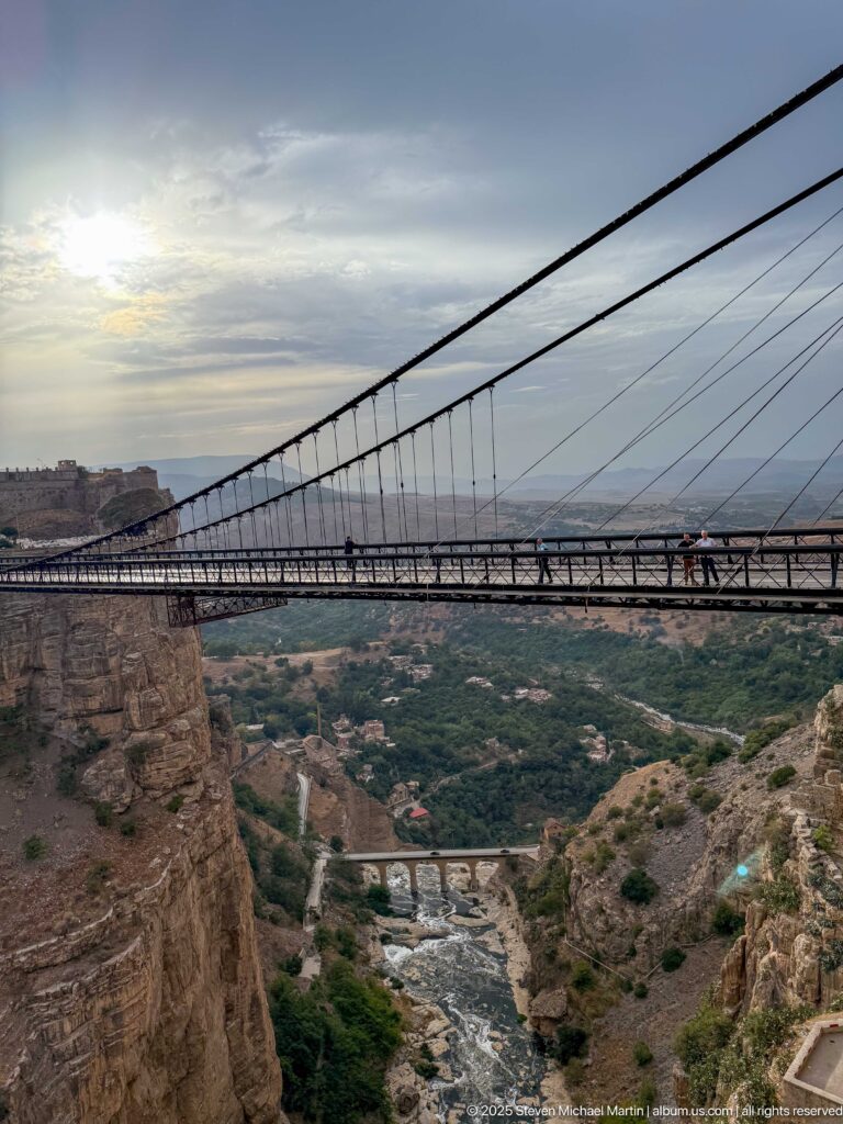 Side view of Sidi M’Cid Bridge deck and suspension hangers high above the Rhumel valley, with pedestrians crossing.