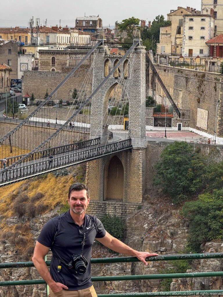 Man at railing in front of Sidi M’Cid Bridge’s stone tower, suspension cables, and metal deck.