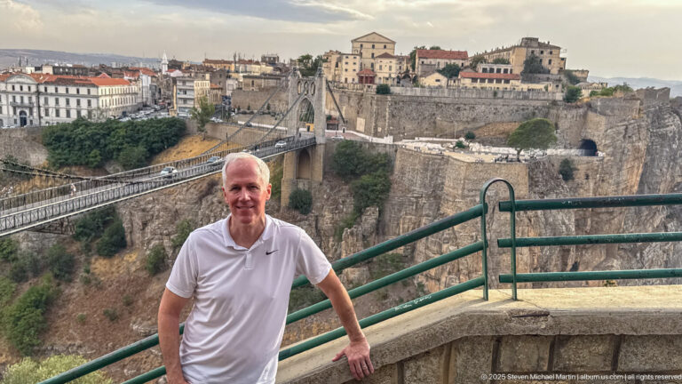 Man at a gorge overlook with Sidi M’Cid Bridge, sheer cliffs, retaining walls, and Constantine buildings behind.