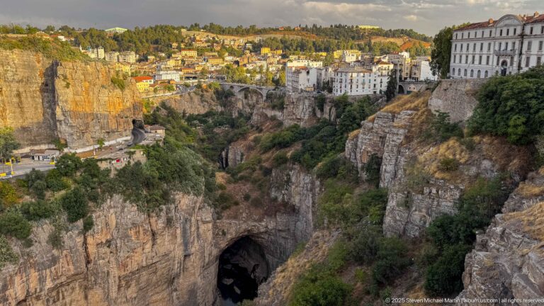 Warm light on Constantine’s cliff-top buildings above a shadowed gorge with a large dark rock opening below.