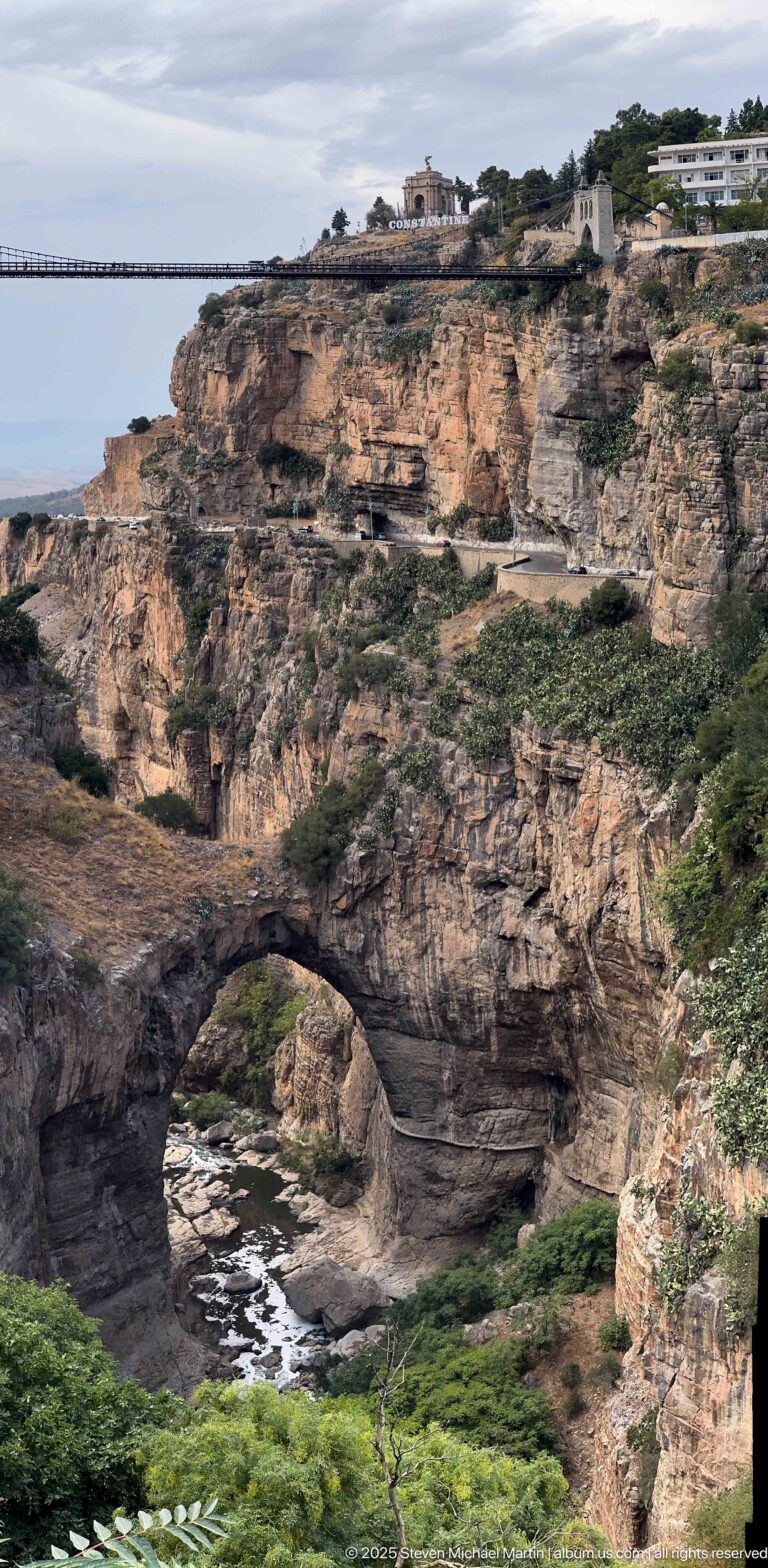 Wide view of a natural rock arch over the Rhumel channel beneath layered cliffs and sparse vegetation.