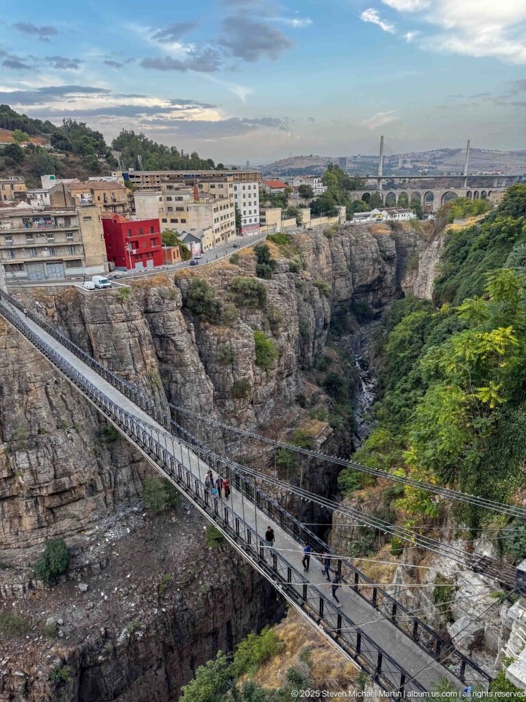 Pedestrians crossing a narrow suspension footbridge above Constantine’s rocky gorge, with city buildings on the far rim.