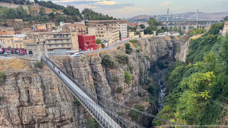 Suspension footbridge crossing a deep gorge toward city buildings, with a larger arched bridge visible in the distance.