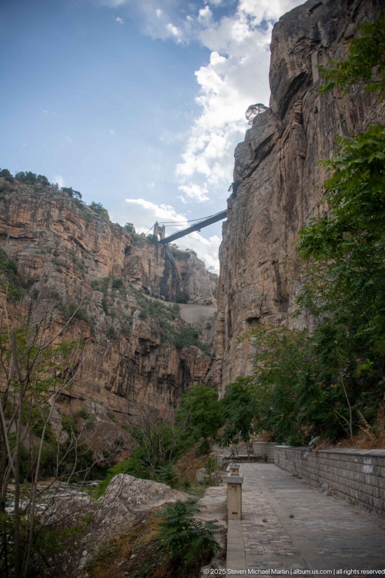 Paved path at the bottom of Constantine’s gorge beneath steep layered cliffs and a high slender bridge.