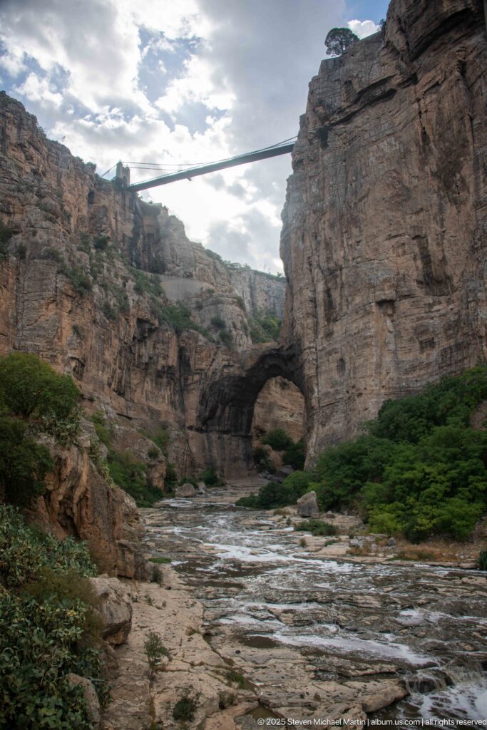 Rocky Rhumel River channel leading toward a large natural arch, with a high bridge spanning the gorge above.