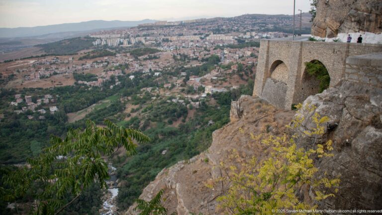Arched stone overlook projecting from a rocky cliff above Constantine, with visitors and the city beyond.