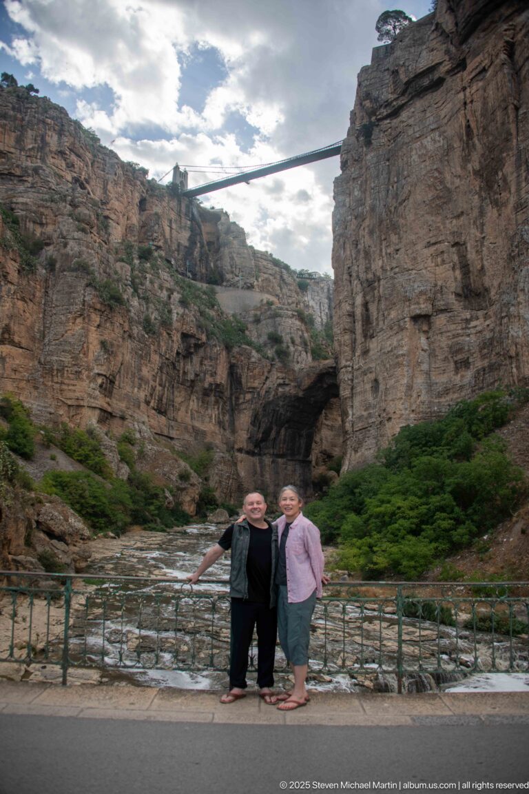Two visitors standing by a railing beside the Rhumel River, with a natural rock arch and high bridge behind.