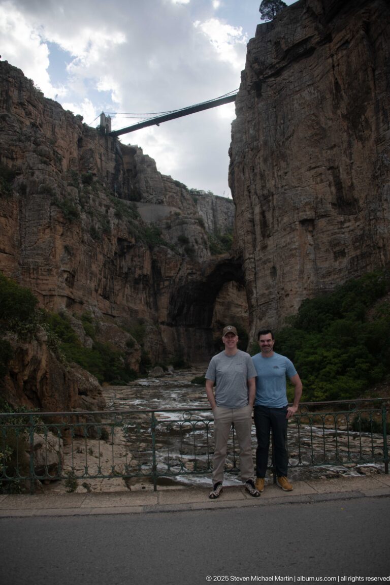 Two visitors posing at the canyon floor before a natural rock opening, with a bridge spanning high overhead.