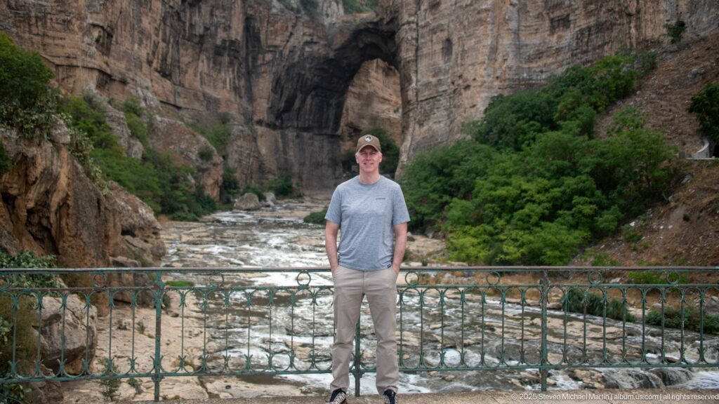 Man standing at a railing above the Rhumel River, with a tall dark natural opening in the gorge behind.