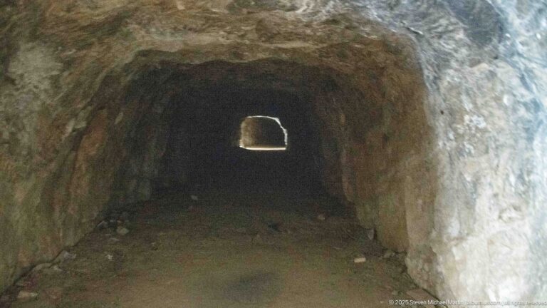 Rough rock-cut tunnel with an uneven floor and a small rectangular opening glowing at the far end.