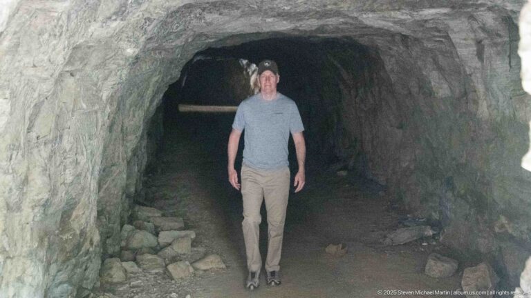 Man standing inside a narrow rough-hewn rock tunnel with loose stones and a dim opening behind him.