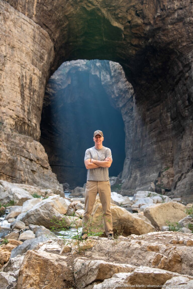 Man with folded arms standing on boulders before a large natural rock arch in Constantine’s gorge.