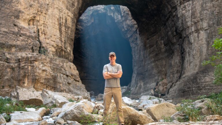 Man standing on boulders before a tall natural gorge opening, with blue light receding through the passage.