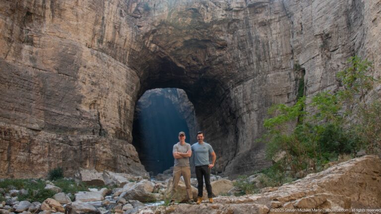Two men standing on rocks before a large natural arch in the layered cliffs of Constantine’s gorge.