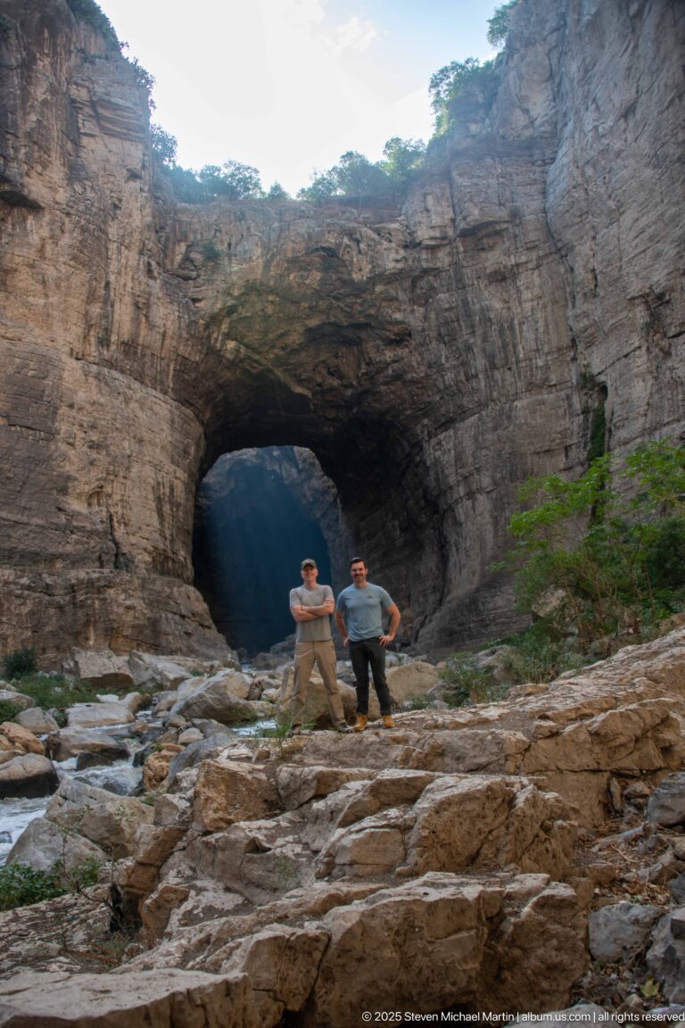 Two men standing beneath a broad natural rock arch, surrounded by layered canyon walls and a rocky riverbed.
