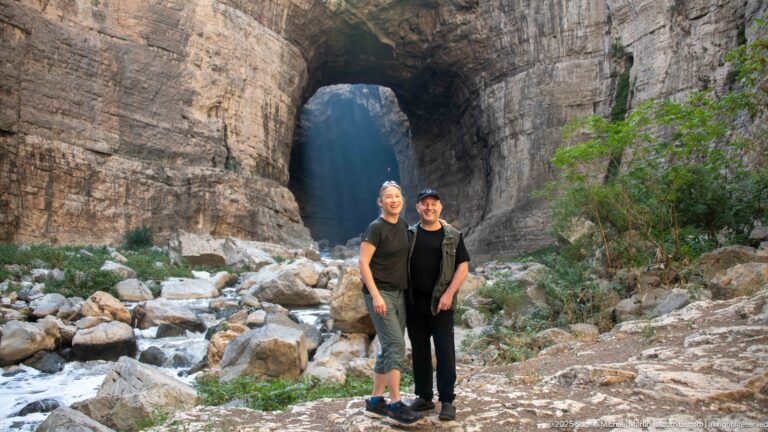 Two visitors standing beside the Rhumel River before a tall natural rock opening in Constantine’s gorge.