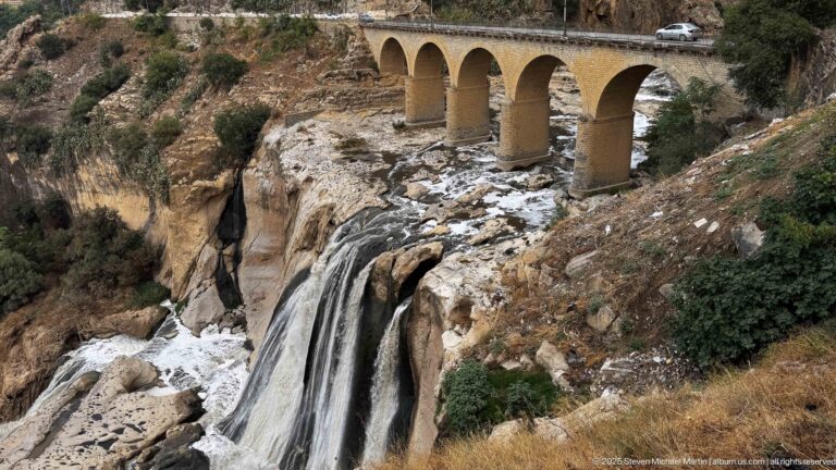 Four-arch masonry bridge above a broad waterfall dropping over pale and dark bedrock in Constantine’s gorge.