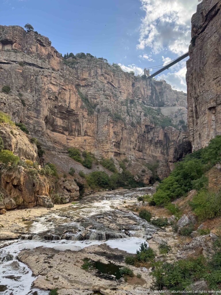 Rocky Rhumel River channel beneath layered gorge walls, with a high bridge and shadowed natural opening above.
