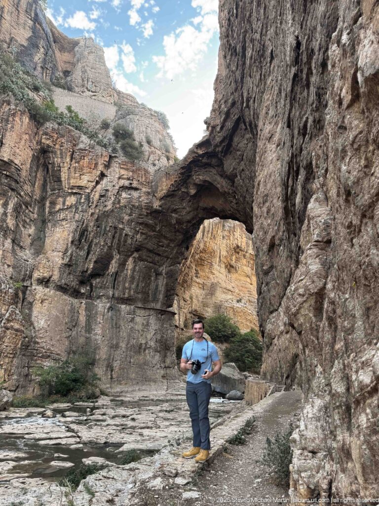 Man standing on rocky ground beside the Rhumel River under a massive natural arch in Constantine’s gorge.