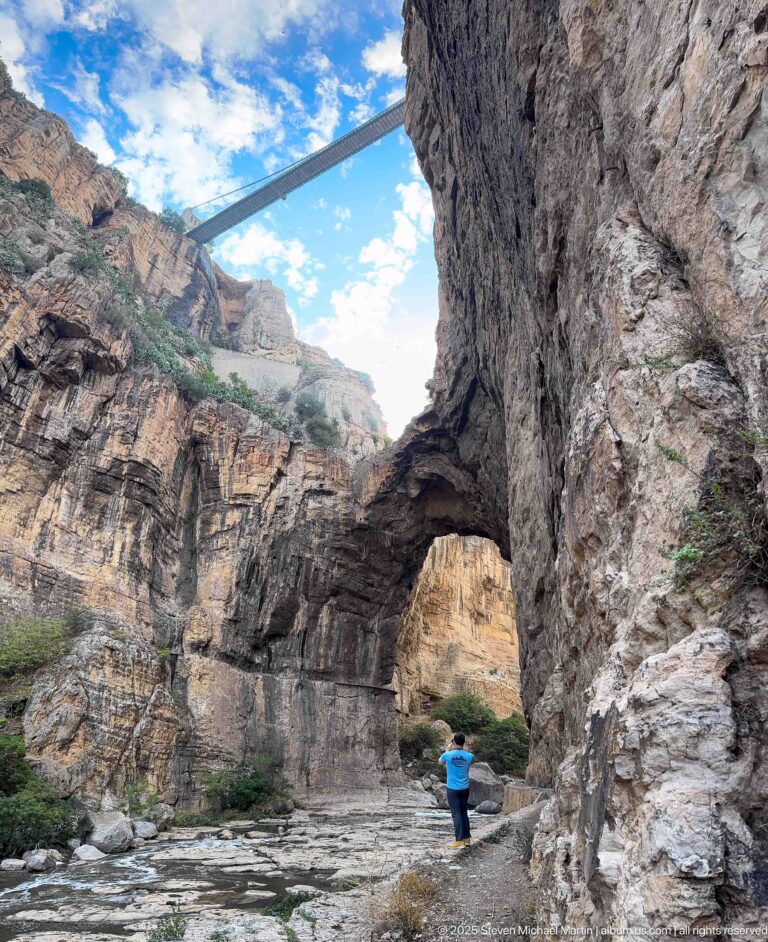 Man beneath a natural rock arch in Constantine’s gorge, with a high bridge spanning the cliffs overhead.