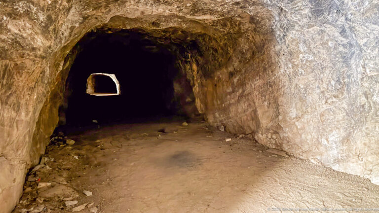Rough rock-cut tunnel with warm stone walls, dirt floor, and small bright opening at the far end.