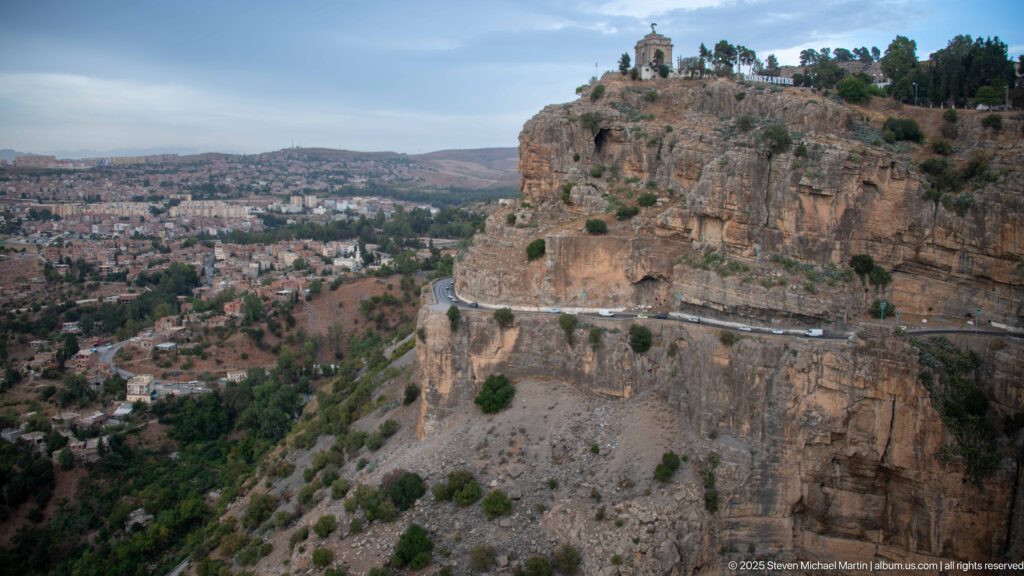 Cliff road curving beneath a rocky escarpment crowned by Constantine’s Monument aux Morts.