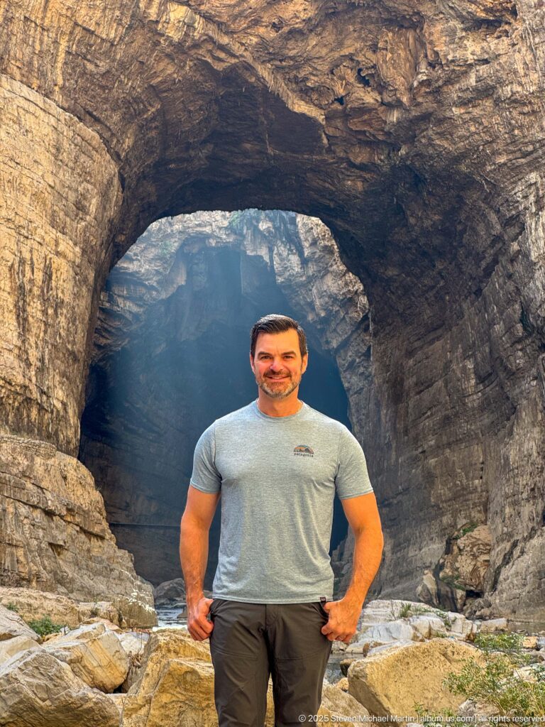 Man standing close to the camera beneath a broad natural rock arch, with boulders and blue-lit gorge behind.