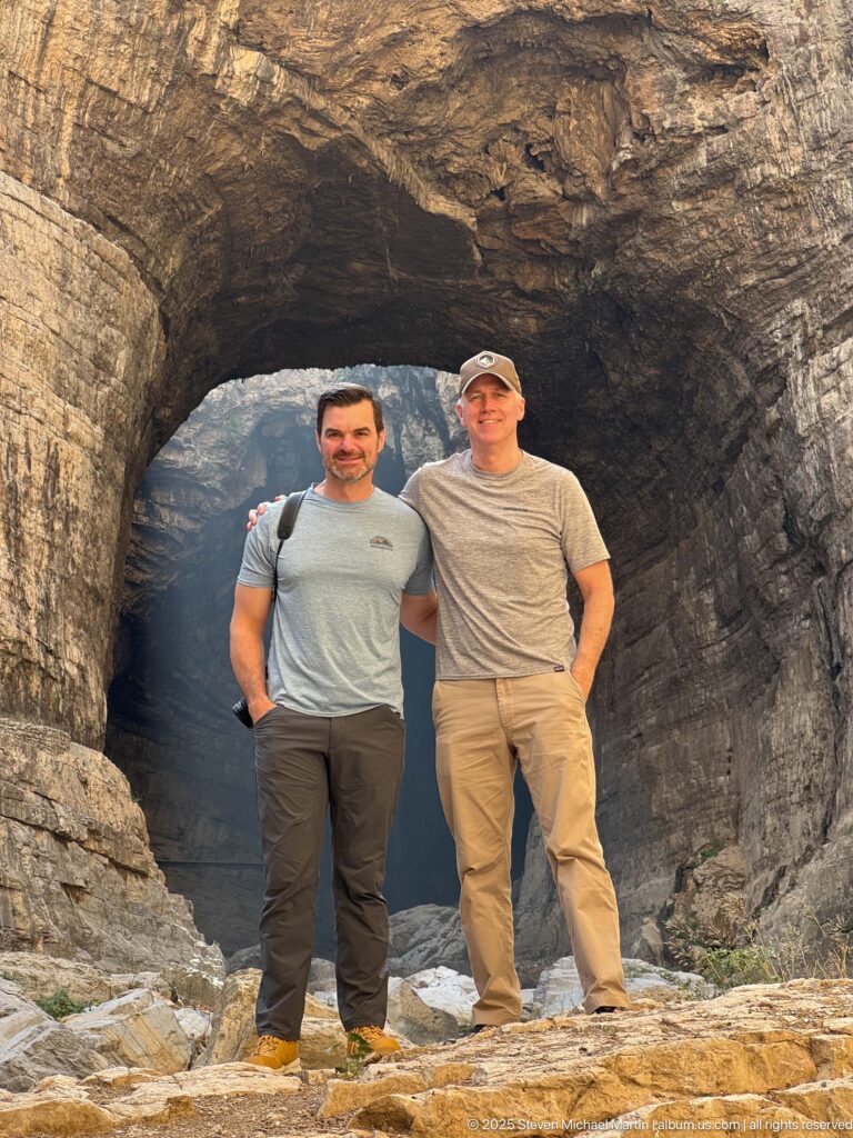 Two men standing together beneath a broad natural rock arch on the rocky floor of Constantine’s gorge.
